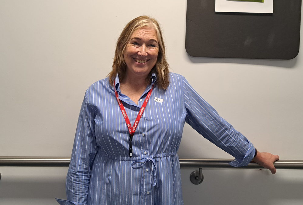 Alison stands smiling at the camera. She is wearing a blue dress and red lanyard.