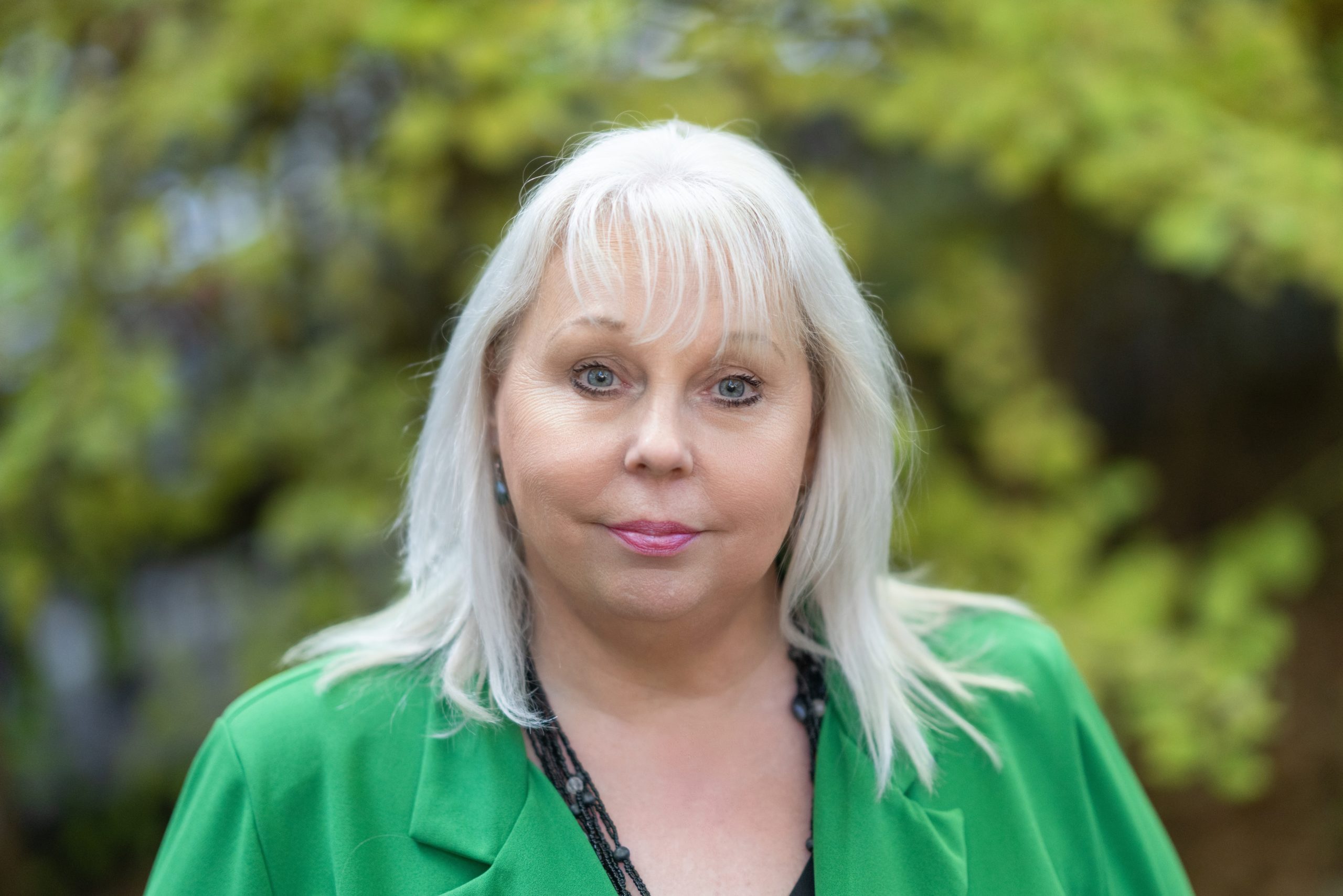 Debbie is smiling at the camera. She has blonde hair and is wearing a bright green blazer, standing in front of a blurred tree in the background.