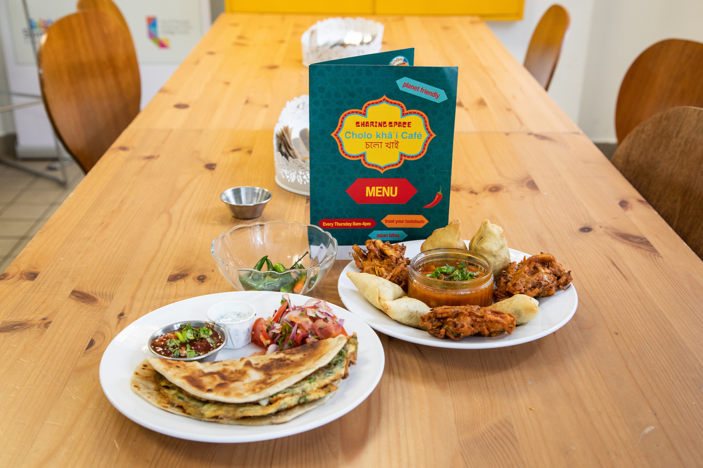 Two plates of Asian food, including samosas and onion bhajis, sit on a wooden table in the café with the menu placed in the middle.