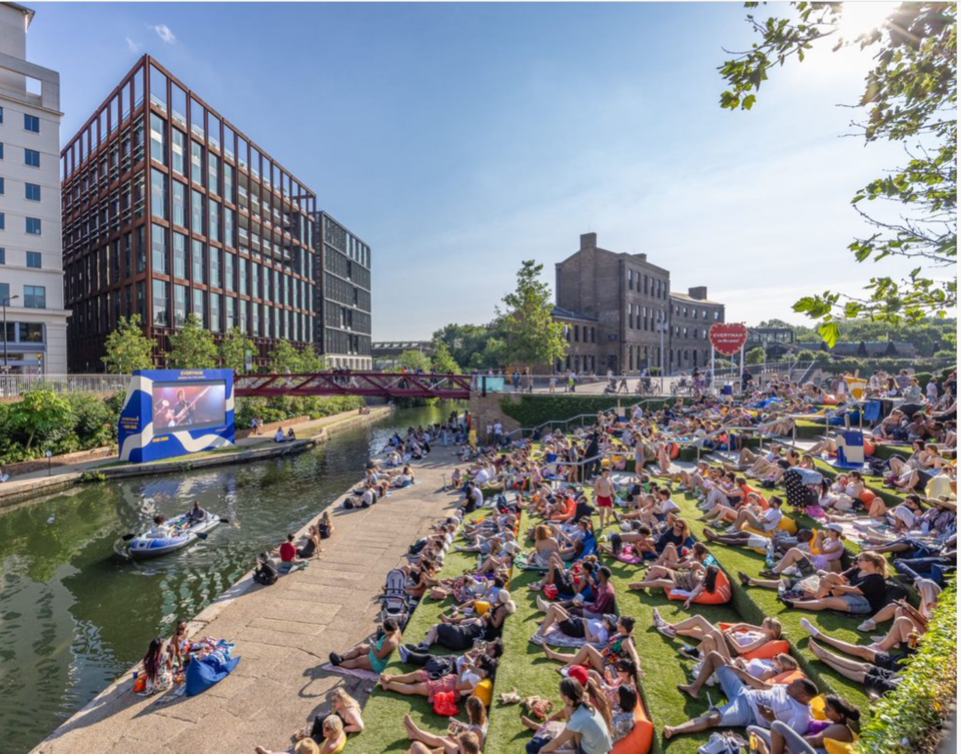 People sat outside in the sun watching a large screen by John Sturrock