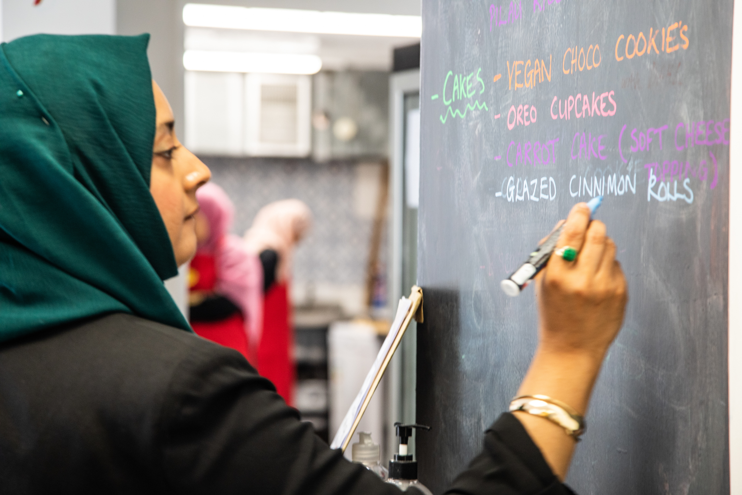 One chef is writing the cakes menu on a blackboard in a chalk pen. She is wearing a forest green hijab and two other chefs are in the kitchen in the background. The menu reads: vegan choco cookies, Oreo cupcakes, carrot cake and glazed cinnamon rolls.