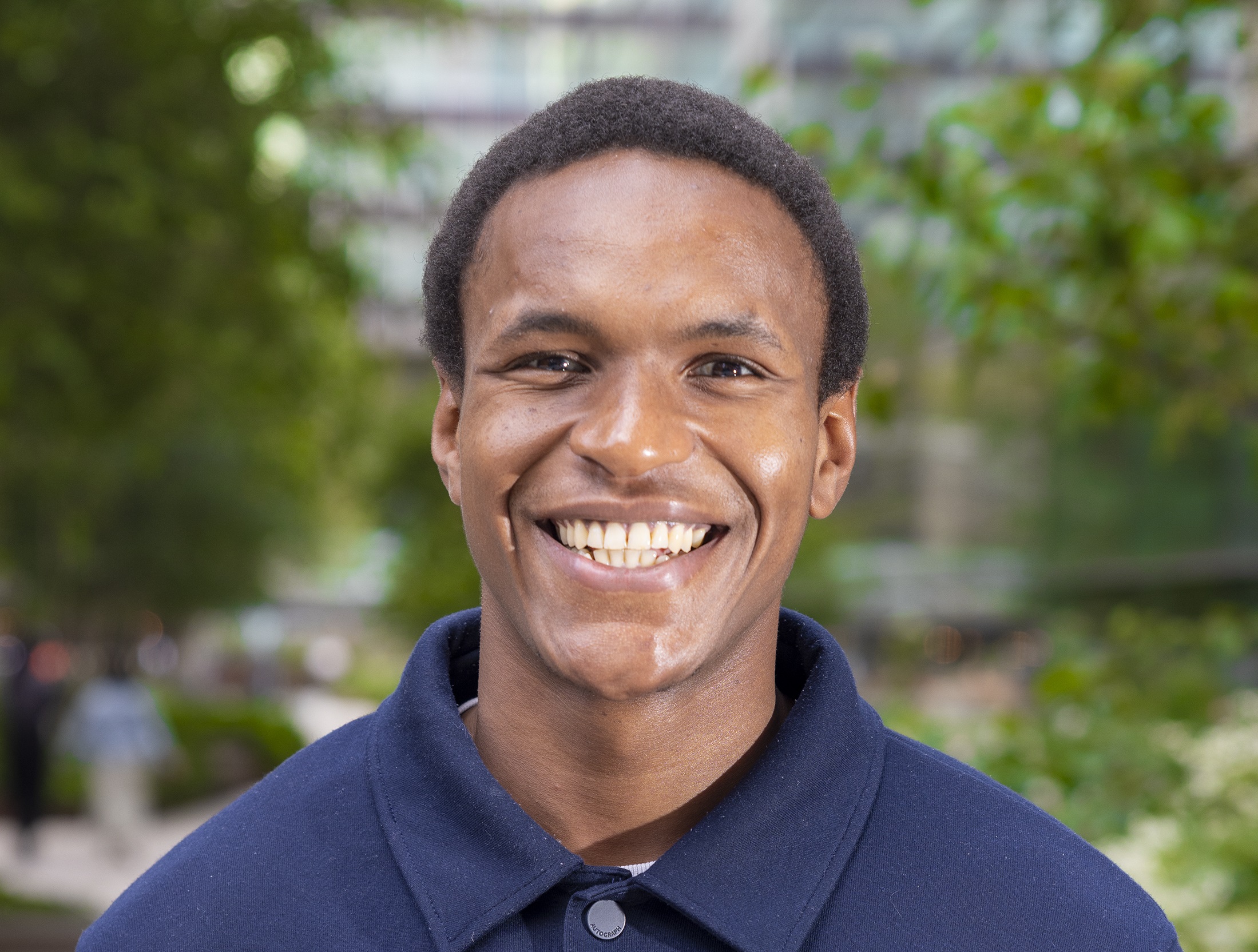 Abraham is smiling at the camera, wearing a navy blue jacket. He is standing in Euston Square, with trees in the background.