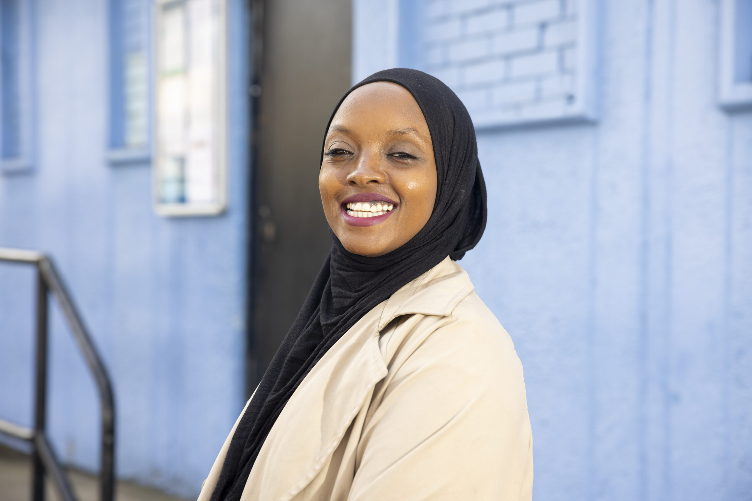 Ikran is smiling directly at the camera. She is wearing a black hijab and cream rain jacket and is standing against the blue wall of a community centre.