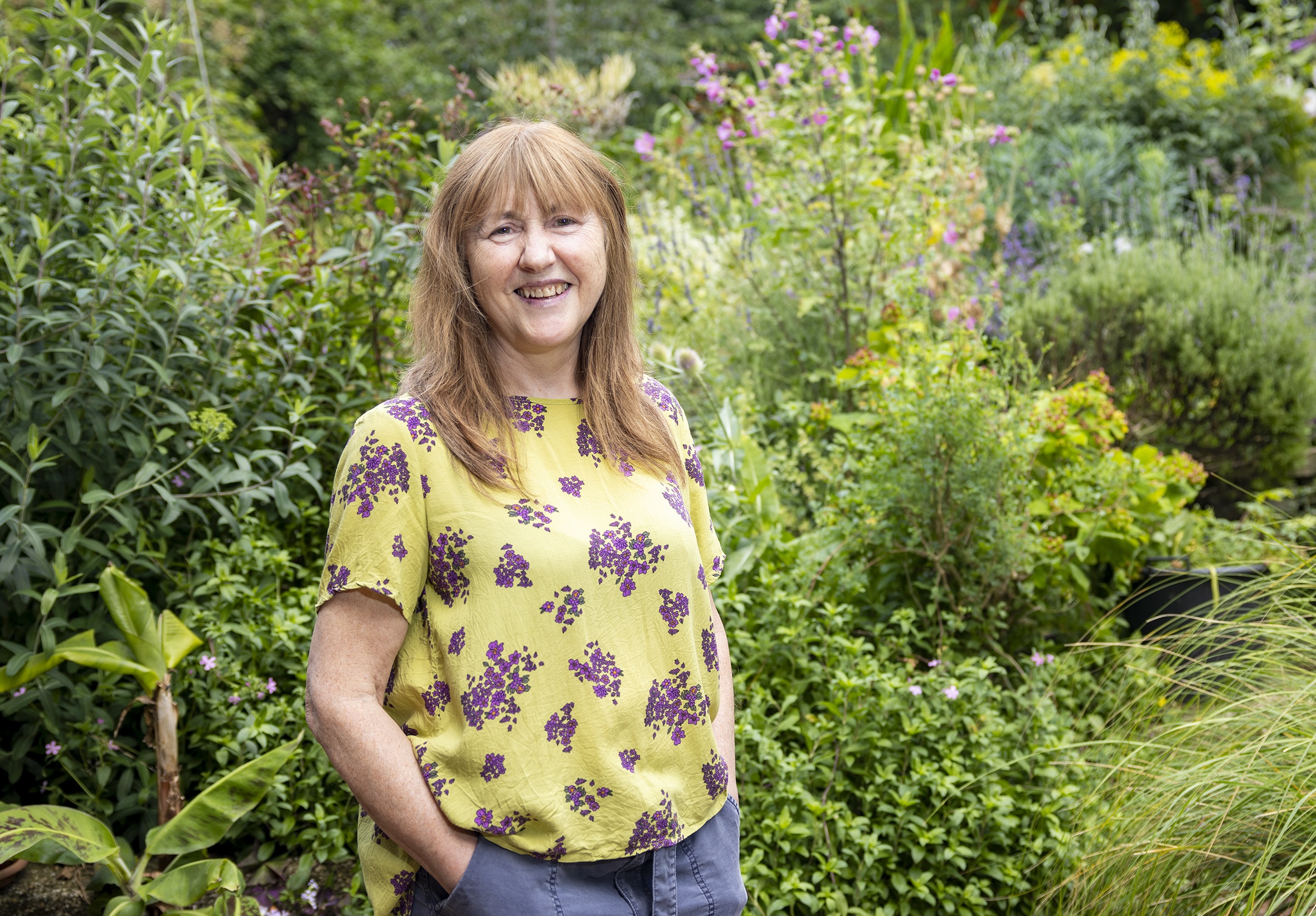 Louise is smiling at the camera, wearing a yellow top with purple flowers on it. She is standing in Phoenix Garden, surrounded by plants.