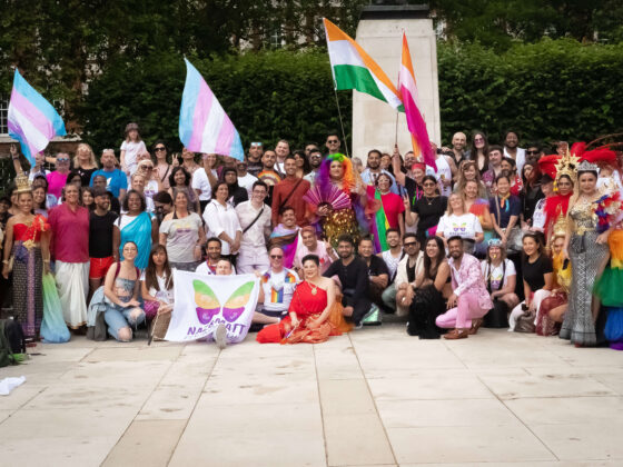 A large group of Naz and Matt Foundation members stand together at London Pride 2023. They are holding LGBTQIA+ flags and banners, smiling at the camera on a sunny day.