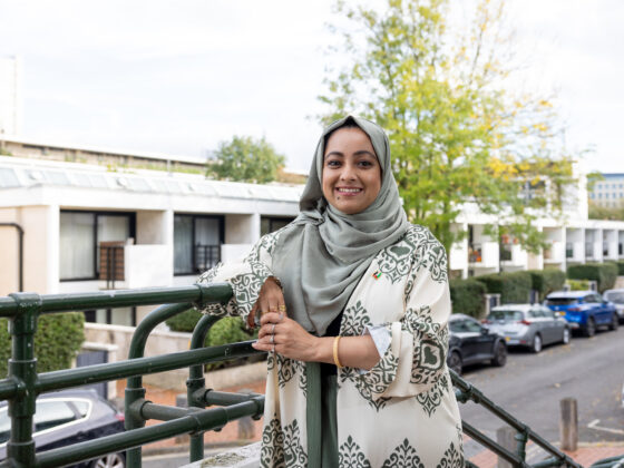 Luthfa is wearing a light green head scarf and cream kaftan with green patterns on it. She is standing in front of one of the blocks on Maiden Lane estate, against green railings – there are trees and cars also in the background. Luthfa is smiling directly at the camera.