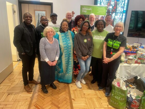 12 Camden Faith representatives at an Interfaith food poverty discussion stand together, alongside food donations. Everyone is posed together and smiling at the camera.