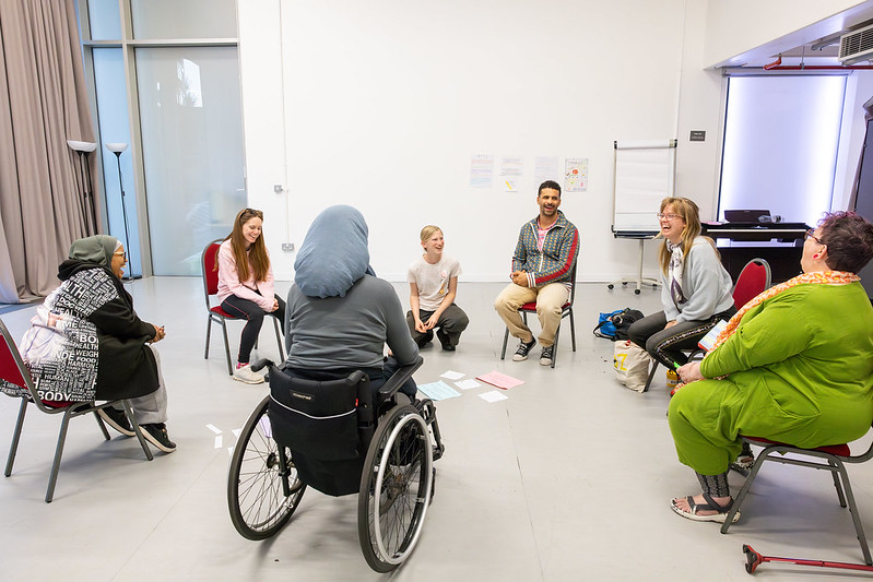 Six participants sit together in a large circle with Nell in a white studio at Old Diorama Arts Centre.