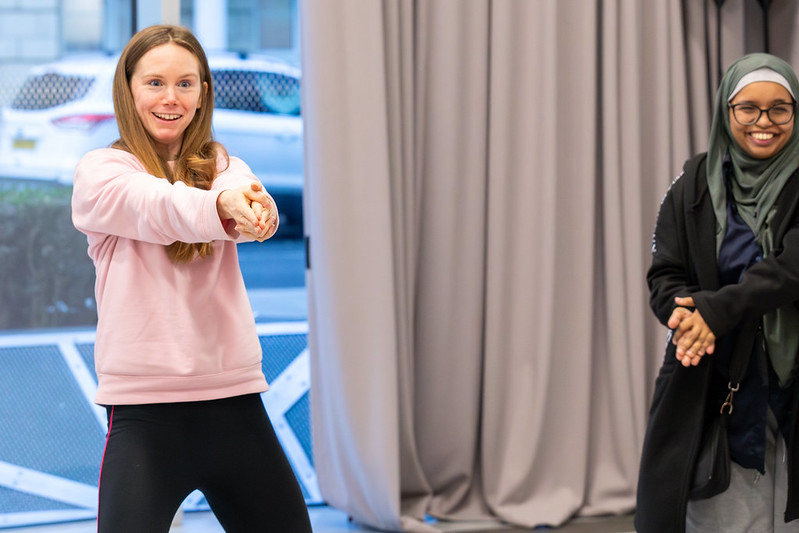 Two participants are playing a group game – the photo is a close-up. One participant is wearing a pink sweatshirt and black leggings, smiling, and the other is wearing a grey hijab, glasses and is smiling.