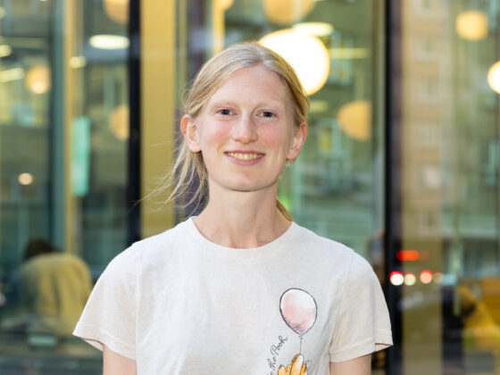 Nell is smiling directly at the camera, standing outside the Old Diorama Arts Centre. She has blonde hair tied in a low ponytail and is wearing a Winnie-the-Pooh t-shirt.