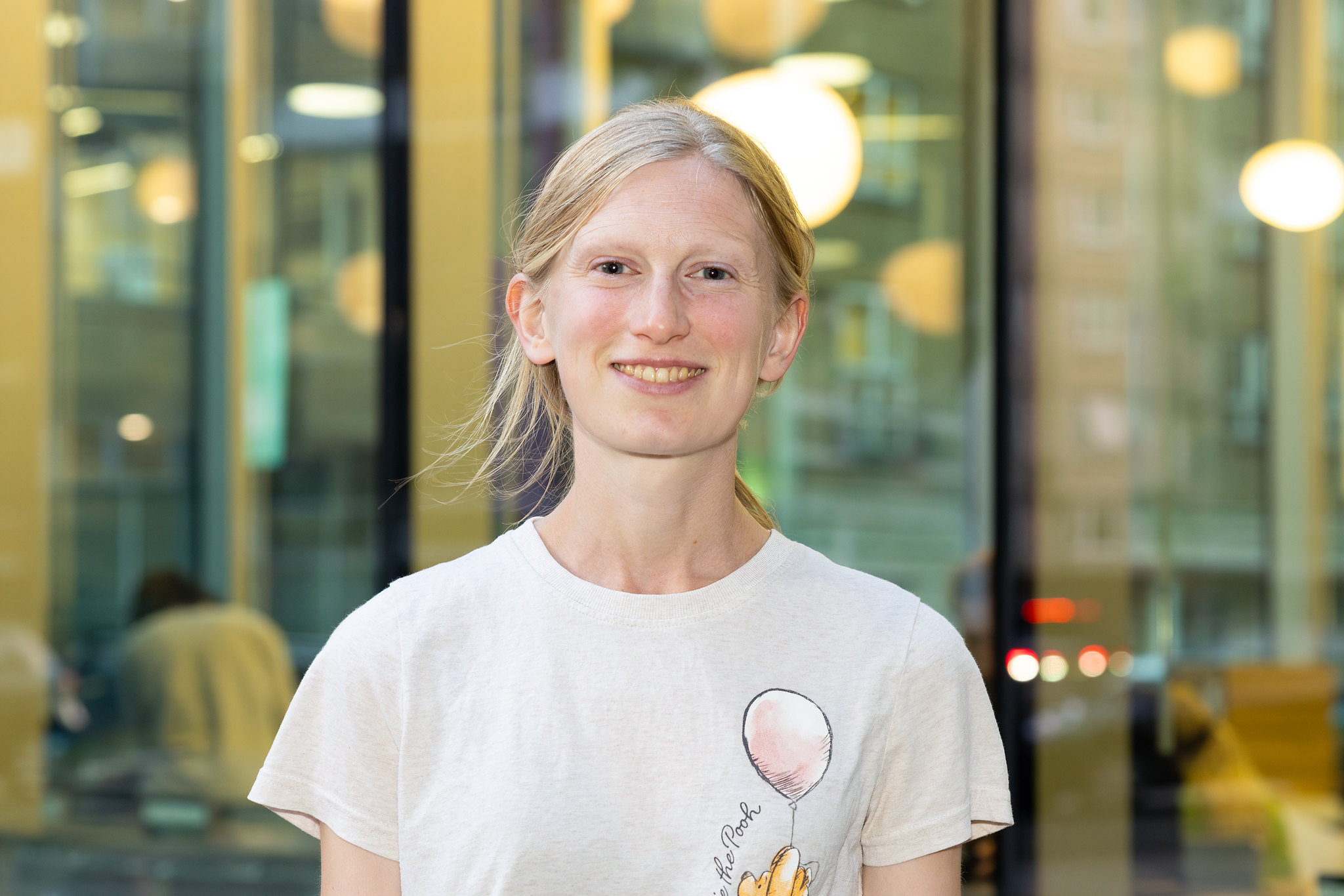 Nell is smiling directly at the camera, standing outside the Old Diorama Arts Centre. She has blonde hair tied in a low ponytail and is wearing a Winnie-the-Pooh t-shirt.