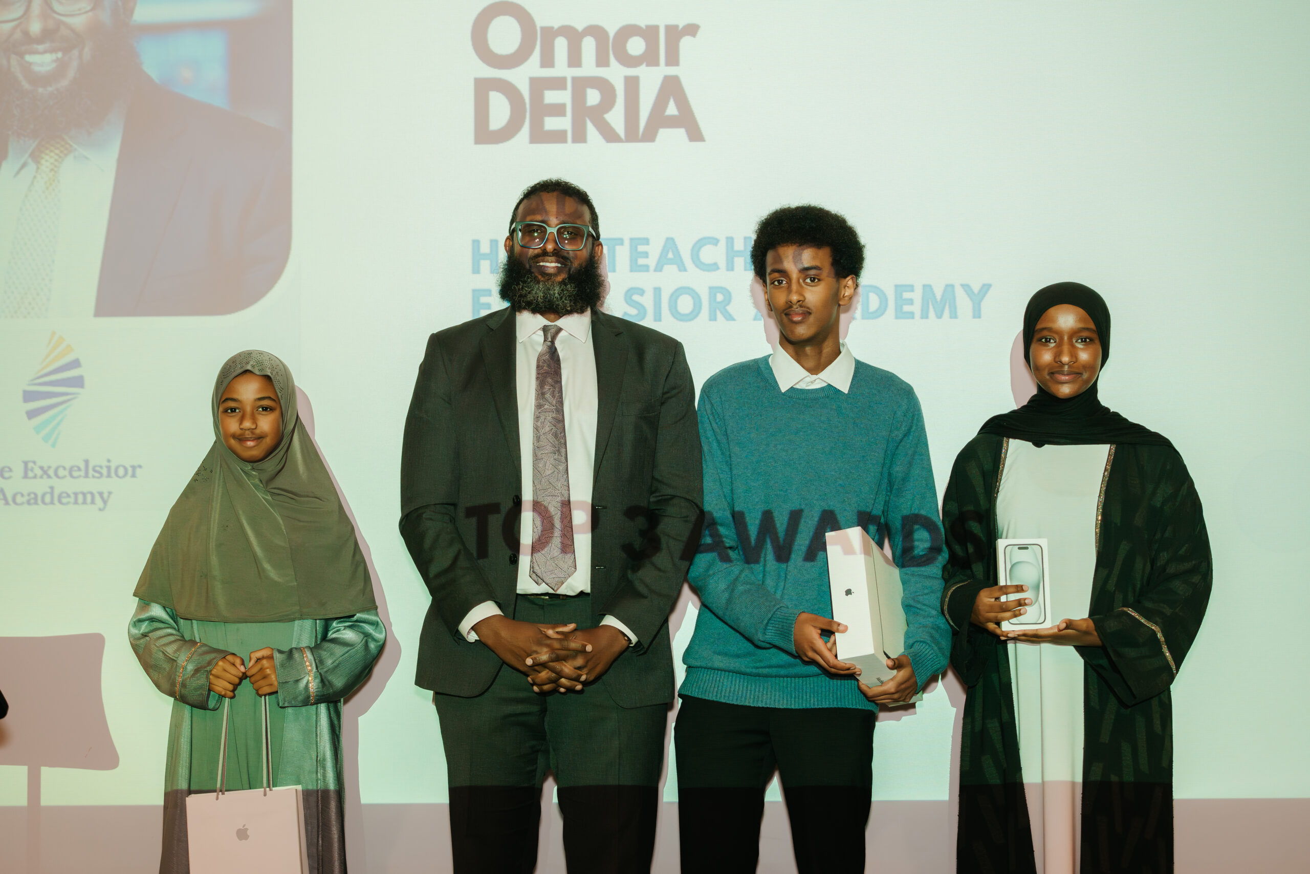 Abdiwahab Ali stands on stage, in line with three young people holding their awards, smiling at the camera.